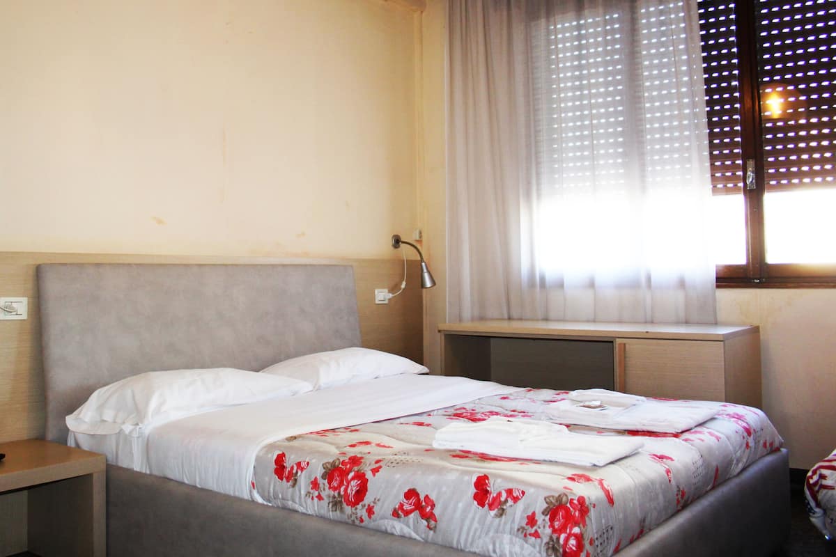 Une chambre d’hôtel avec un lit, des tables de chevet, une lampe et une fenêtre avec des stores.