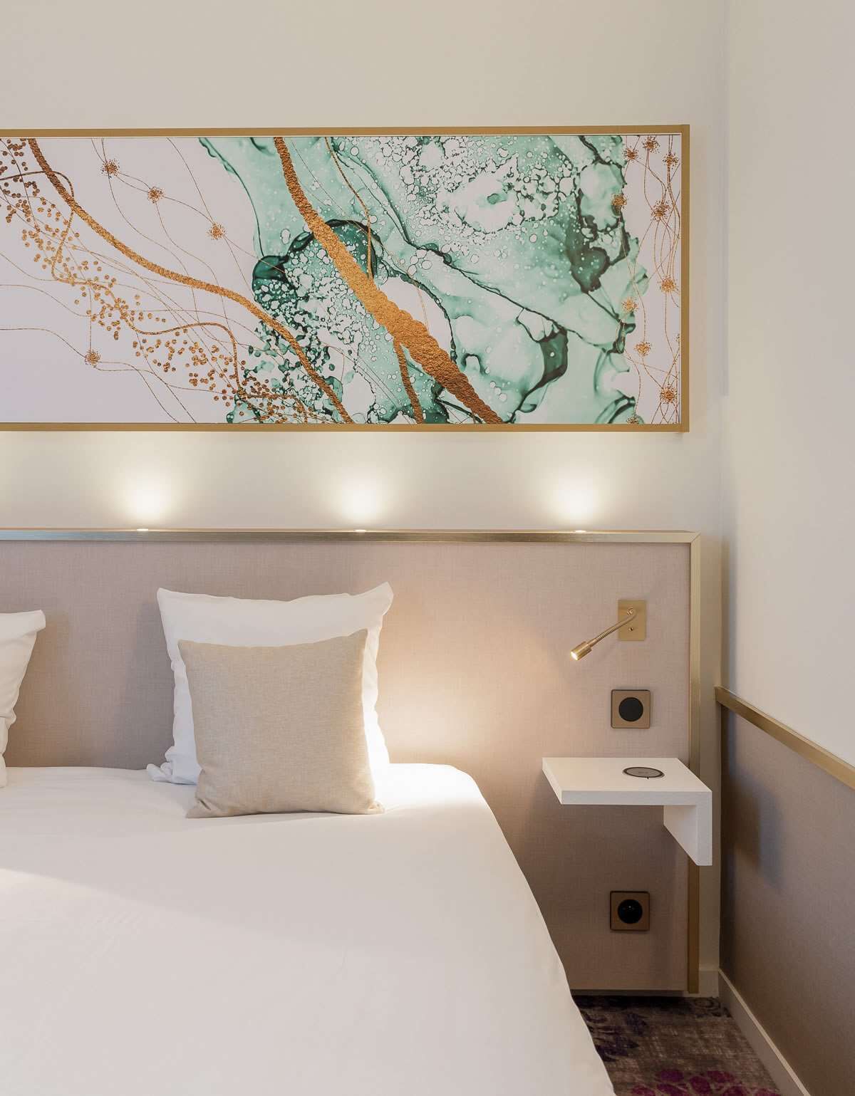 Et hotelværelse med et stort abstrakt maleri over sengen, et natbord med en lampe, og en lille hylde.
