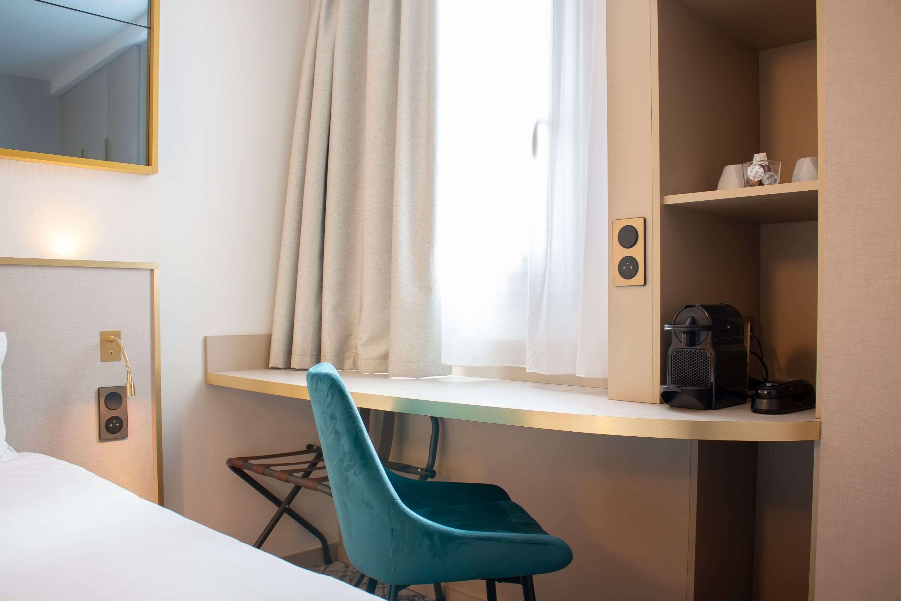 Hotelkamer met een bed, een bureau met stoel, een raam met gordijnen, een koffieapparaat en een plank met spullen.