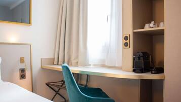 Hotelkamer met een bed, een bureau met stoel, een raam met gordijnen, een koffieapparaat en een plank met spullen.
