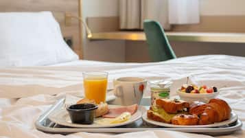 Bandeja de desayuno con cruasanes, jamón, tostadas, un tazón de fruta, un vaso de jugo de naranja, una taza de café y un recipiente pequeño de yogur sobre una cama.