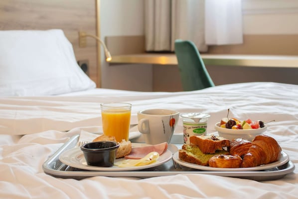 Bandeja de desayuno con cruasanes, jamón, tostadas, un tazón de fruta, un vaso de jugo de naranja, una taza de café y un recipiente pequeño de yogur sobre una cama.