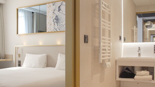 Habitación de hotel con cama, un espejo grande, y un baño blanco con lavamanos y toallero.
