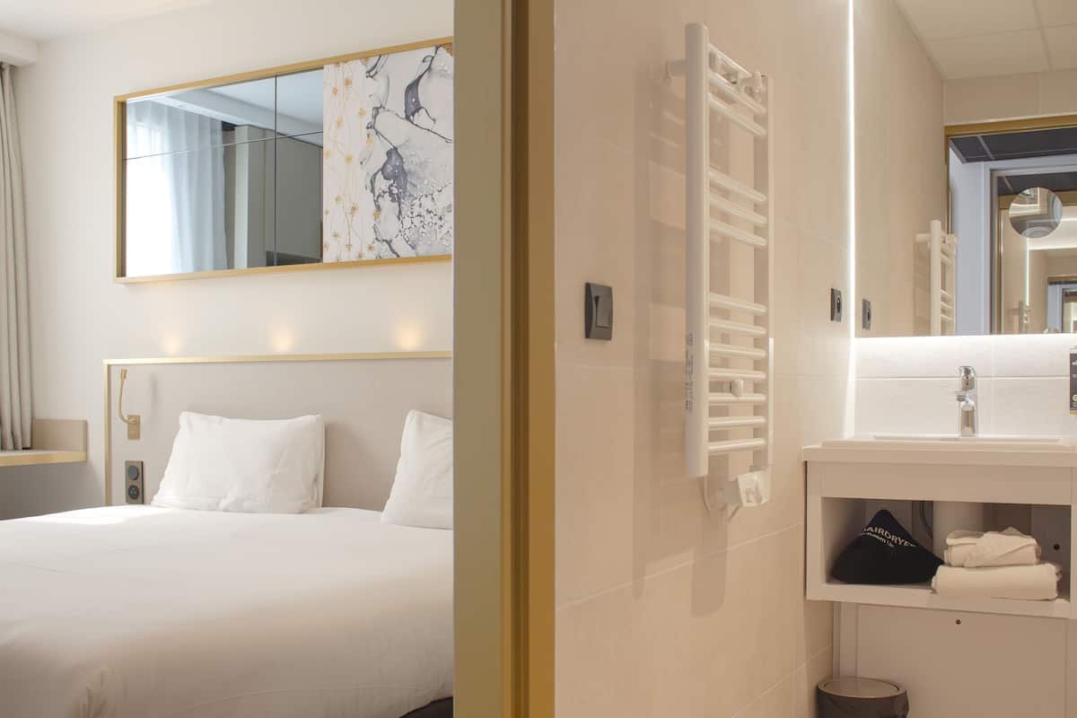 Une chambre d’hôtel avec un lit, un grand miroir, une salle de bain blanche avec un lavabo et un porte-serviettes.
