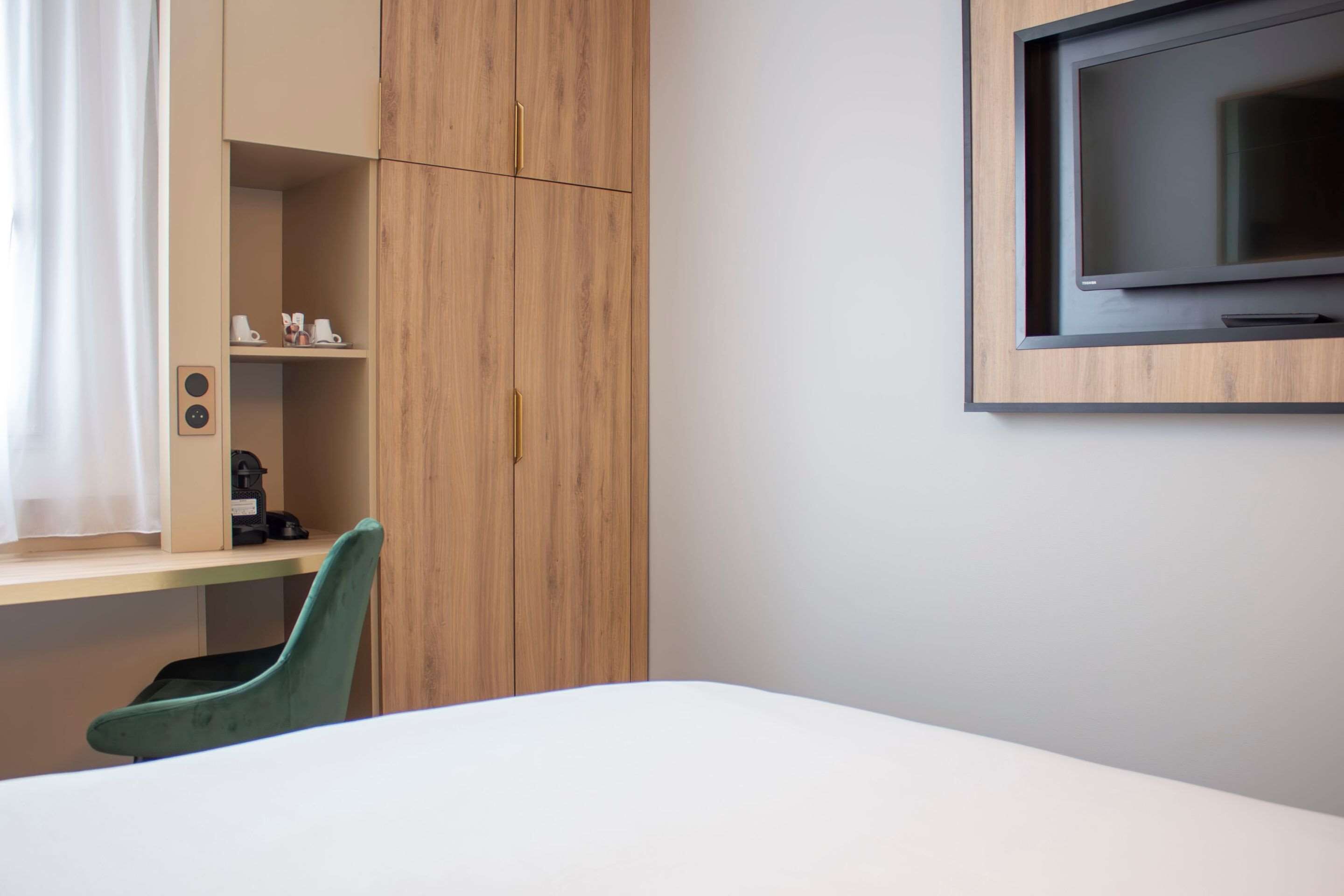 A hotel room with a bed, a desk, a chair, a TV mounted on the wall, and a wardrobe with a mirror.