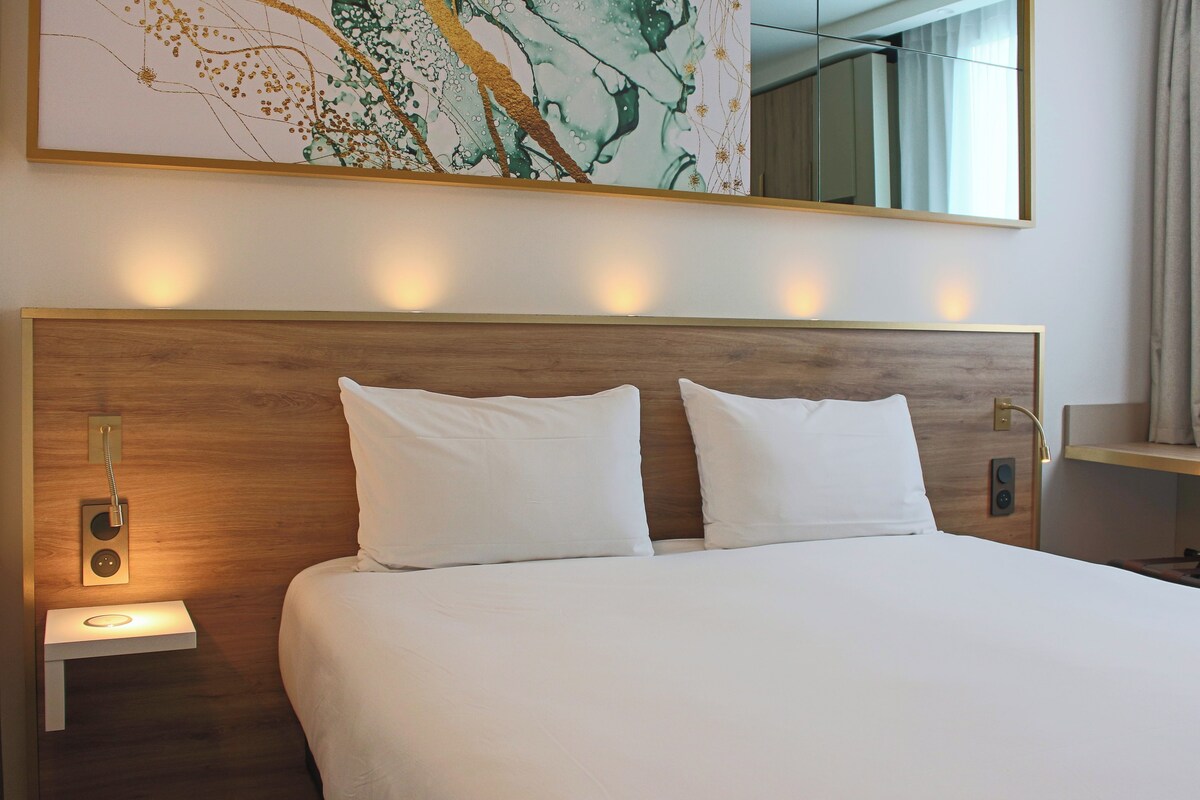 Une chambre d’hôtel avec un grand lit, deux tables de chevet, une lampe fixée au mur et un grand tableau abstrait au-dessus du lit.