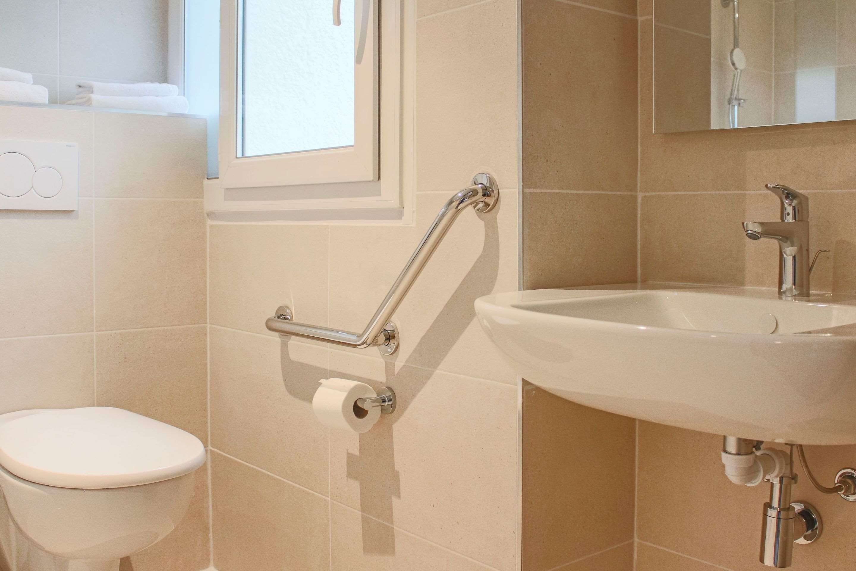 Une salle de bain équipée de toilettes, d’un lavabo, d’une barre d’appui et d’une fenêtre.