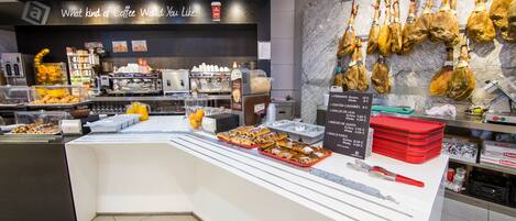 A coffee shop counter with pastries, a menu board, and hanging cured meats.