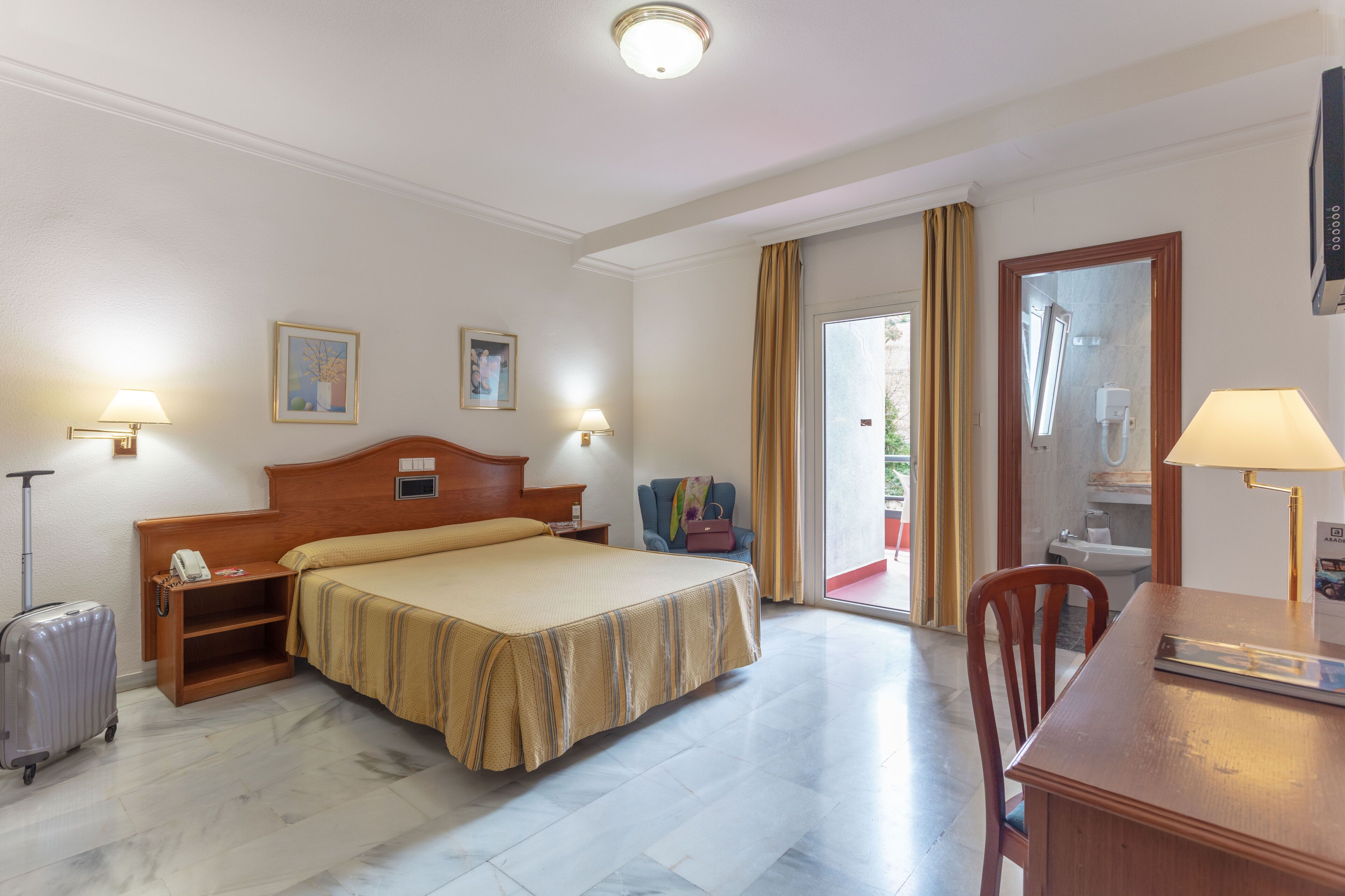 Habitación de hotel con cama, escritorio, silla, maleta y ventana con cortinas.