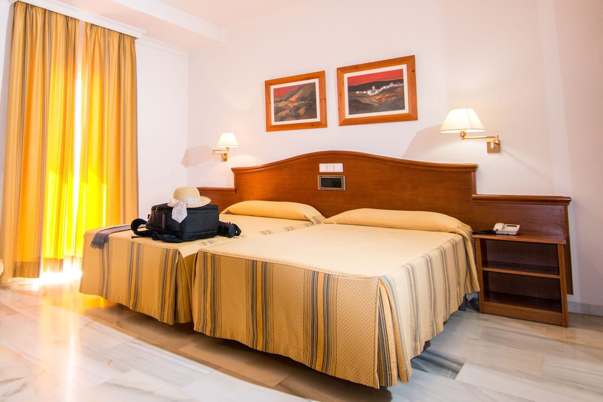 Ein Hotelzimmer mit einem Bett, einem Nachttisch, einer Lampe, einem Koffer und einem Hut auf dem Bett.