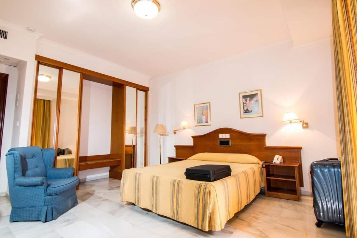 Ein Hotelzimmer mit einem Bett, einem blauen Sessel, einem Nachttisch mit Lampe und einem Koffer.