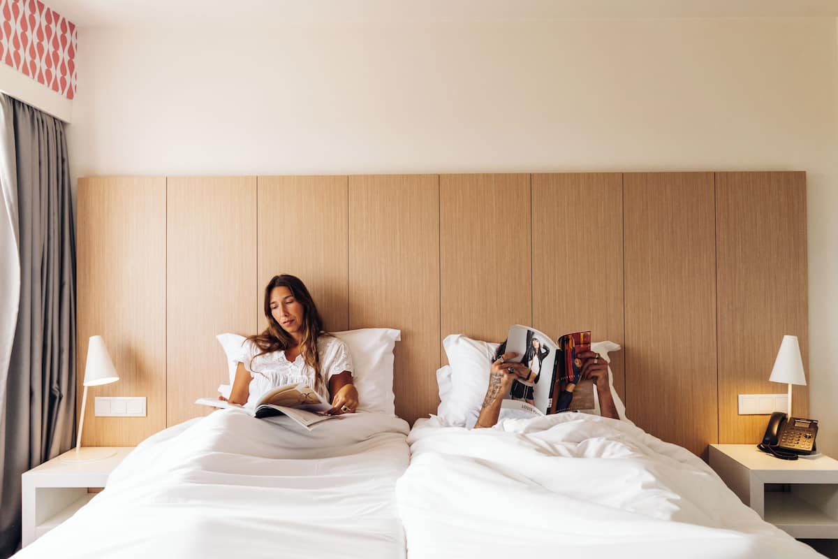 Habitación de hotel con cabecera de madera, ropa de cama blanca, lámpara de noche y teléfono en la mesita de noche.