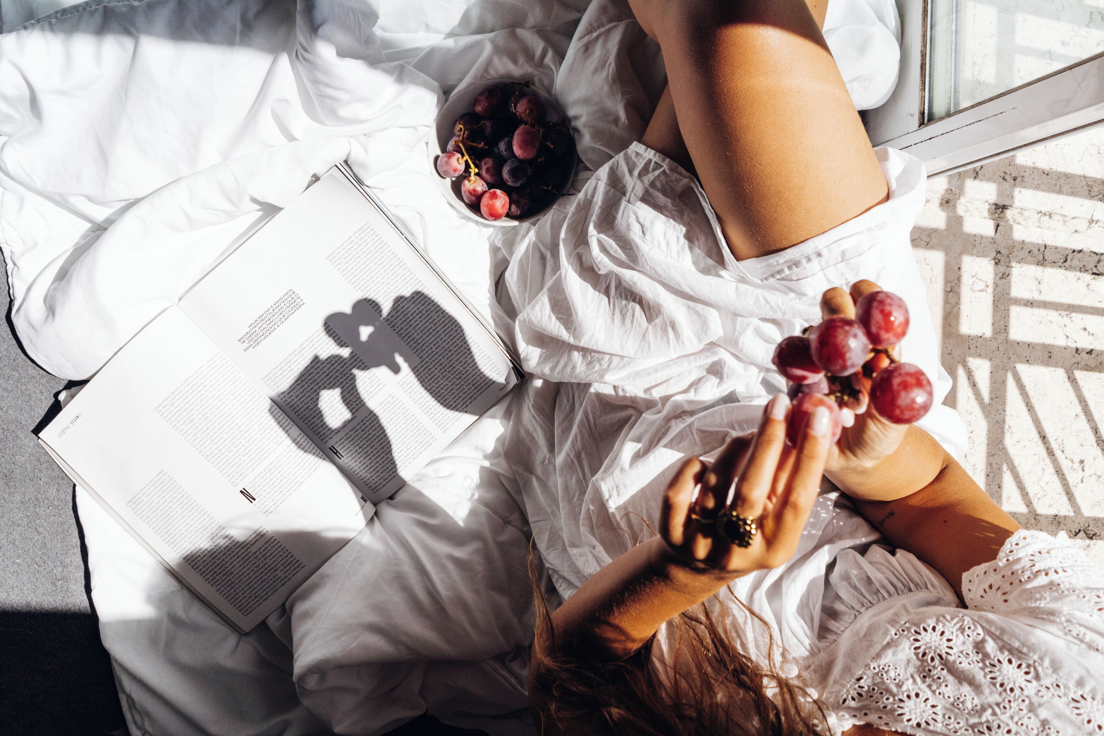 Une personne allongée sur un lit, un livre à la main et un bol de raisins à côté d’elle.