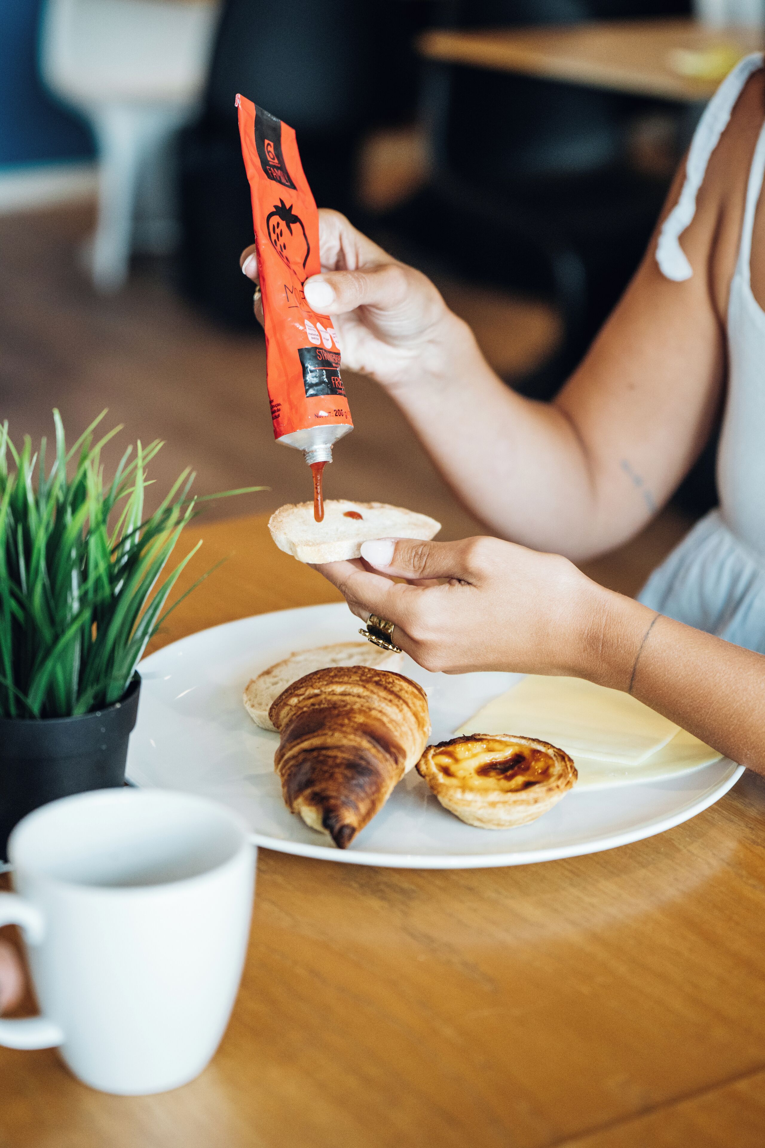 Una persona sosteniendo un tubo de mermelada, extendiéndola sobre un croissant en un plato con una taza y una pequeña planta.