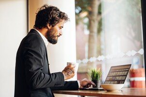 Egy fekete öltönyt viselő személy ül egy asztalnál, előtte egy laptop és egy kávéscsésze van.