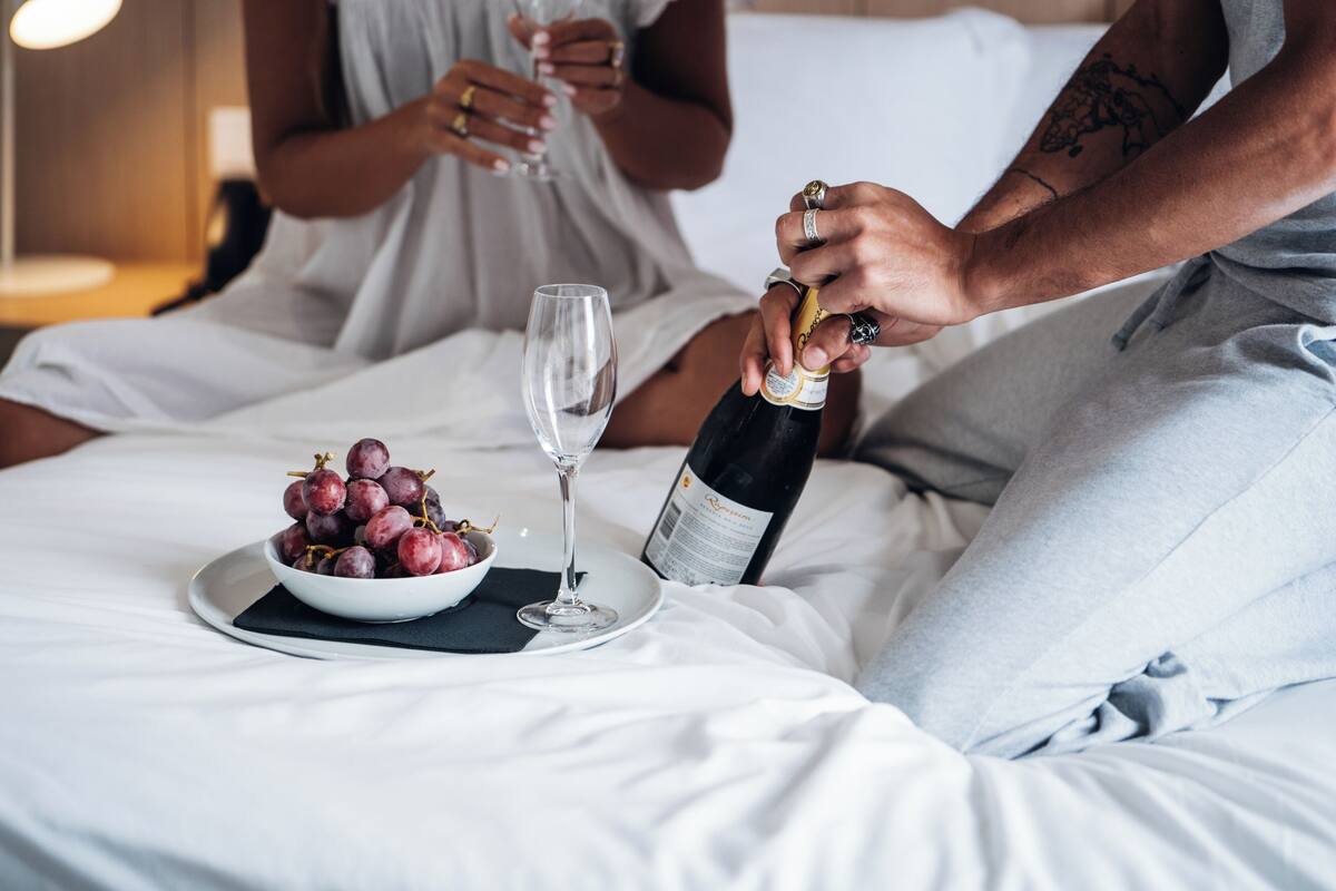 Una cama con un bol de uvas y una copa de vino, una botella de champán siendo abierta, y una persona sirviendo champán en la copa.