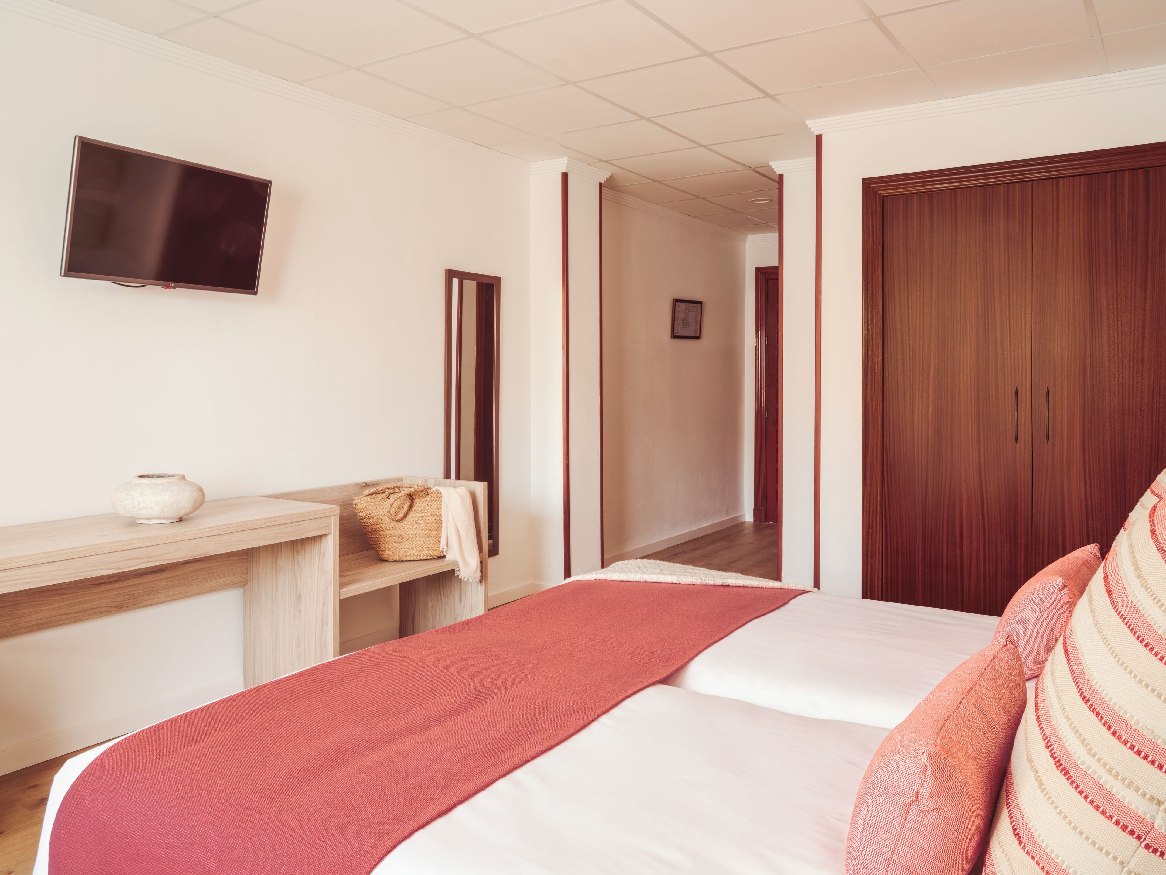 Habitación de hotel con cama, televisor montado en la pared, armario de madera y una pequeña estantería con un jarrón y una cesta.