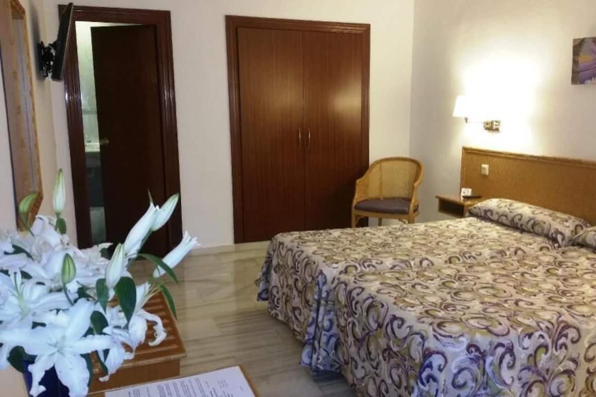 Une chambre d’hôtel avec un lit, une chaise, une armoire en bois et une lampe fixée au mur.
