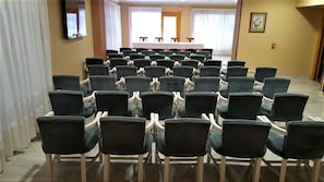 Un salón de conferencias con filas de sillas orientadas hacia una larga mesa, y una pantalla de proyector al frente.