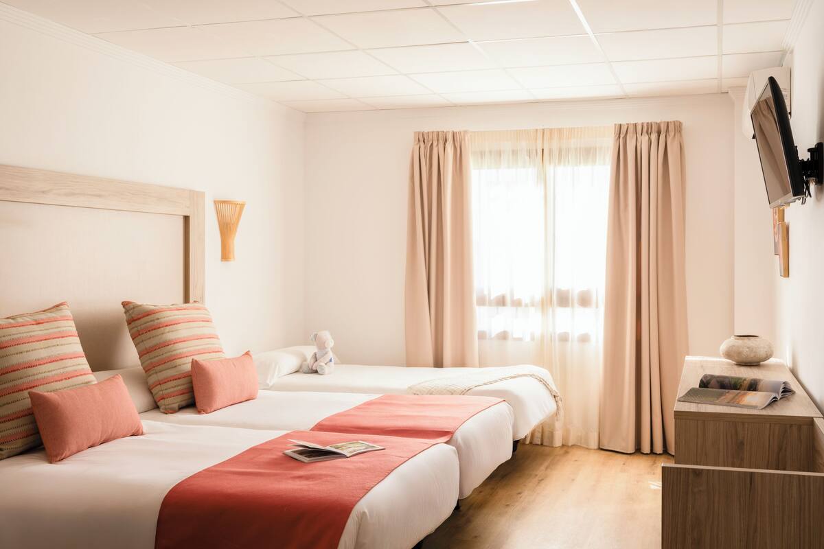 Une chambre d’hôtel avec deux lits, un bureau en bois, un vase et un téléviseur fixé au mur.