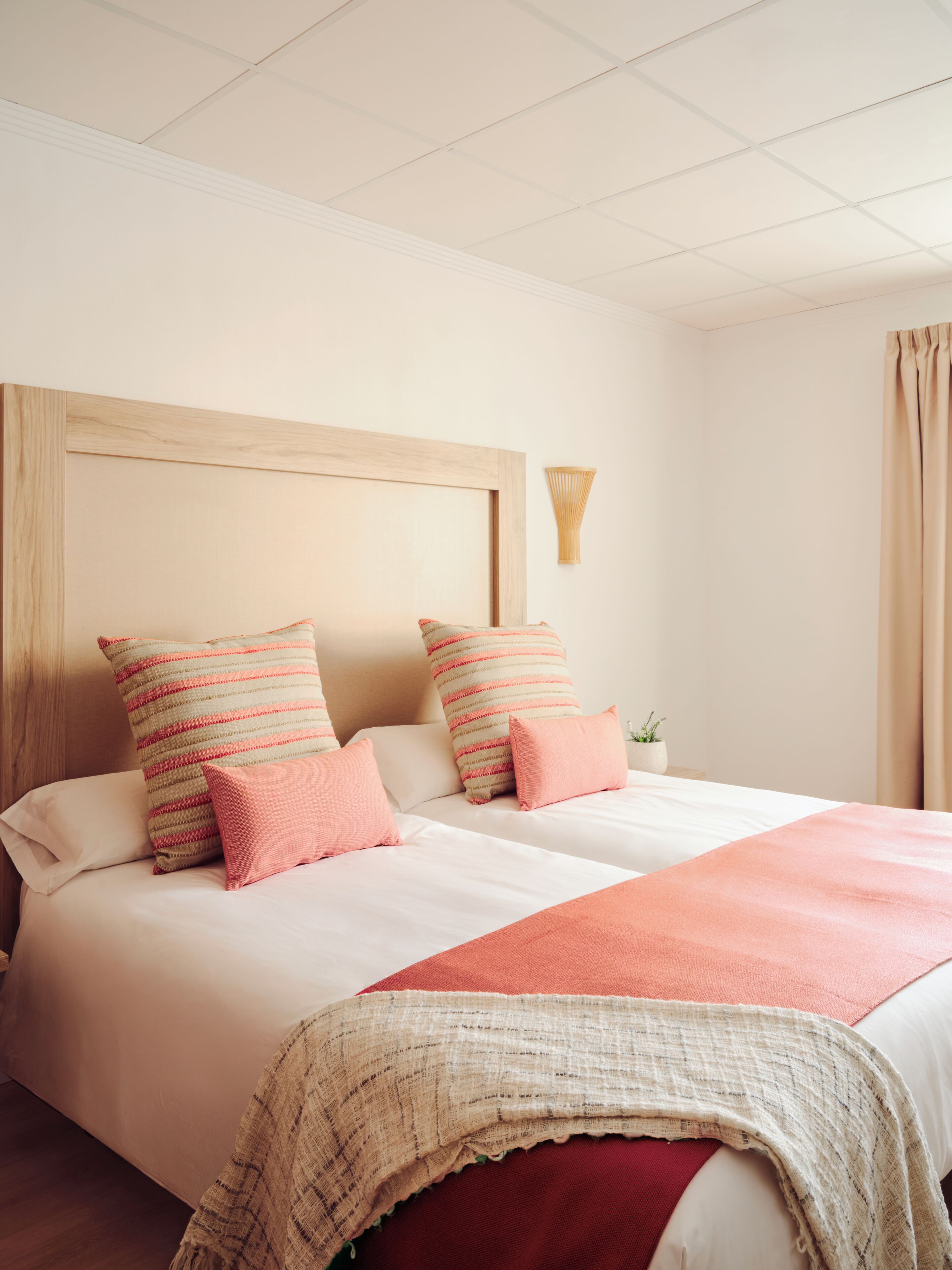 Habitación de hotel con una cama grande, dos almohadas, una mesita de noche con una planta y una ventana con cortinas.