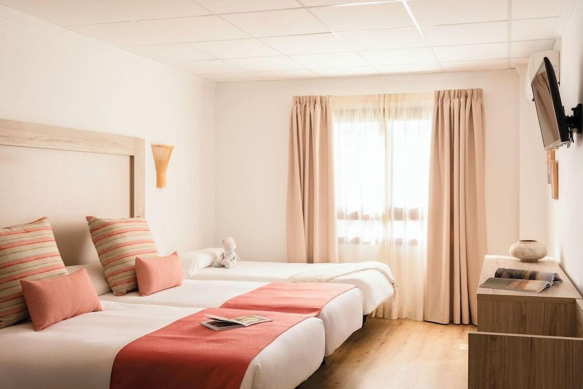 Una habitación de hotel con dos camas, un cabecero de madera, una mesita de noche con un jarrón y un televisor montado en la pared.
