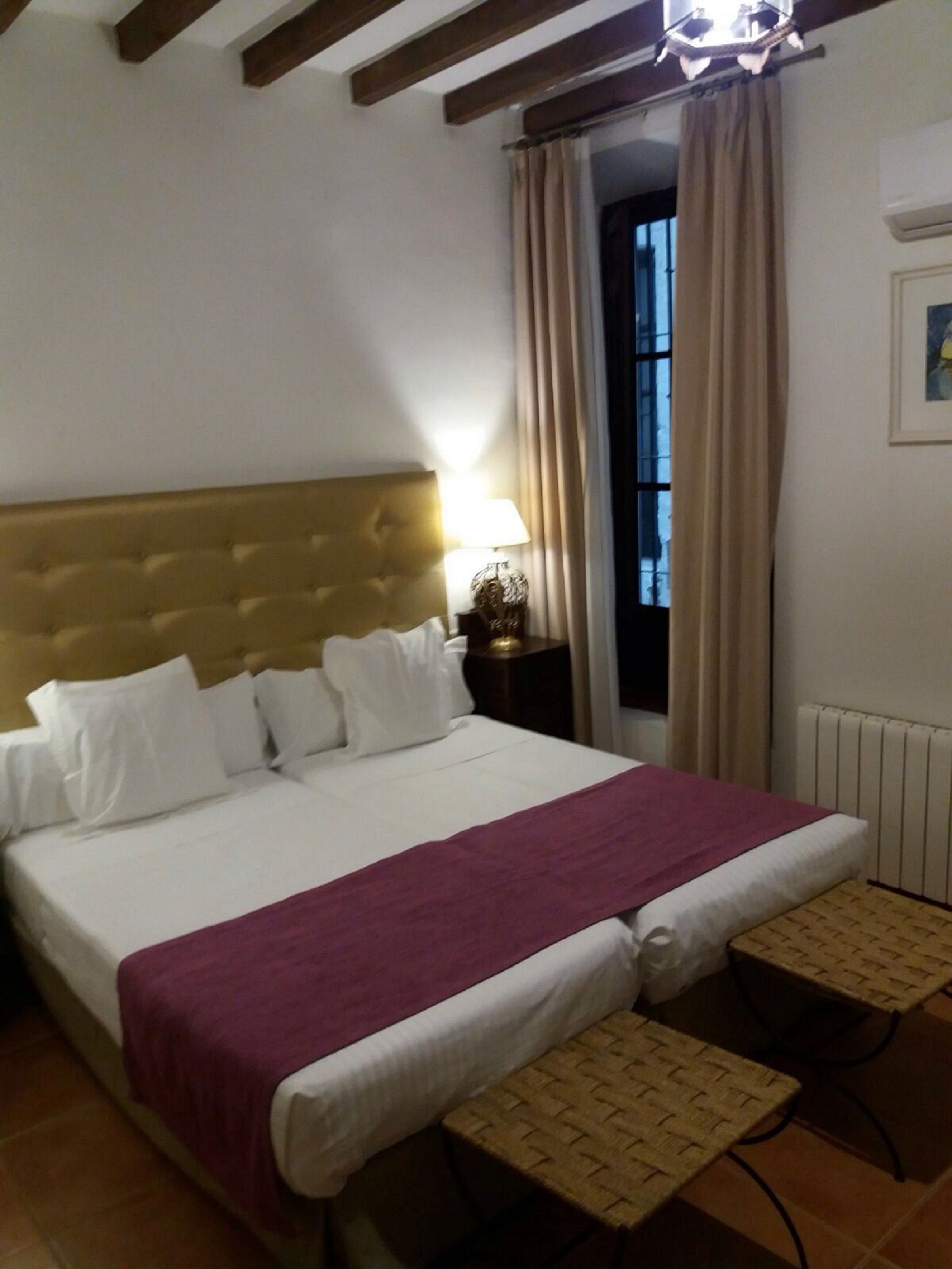 A hotel room with a large bed, a headboard, a bedside table with a lamp, a window with curtains, and a small table with a woven tablecloth.