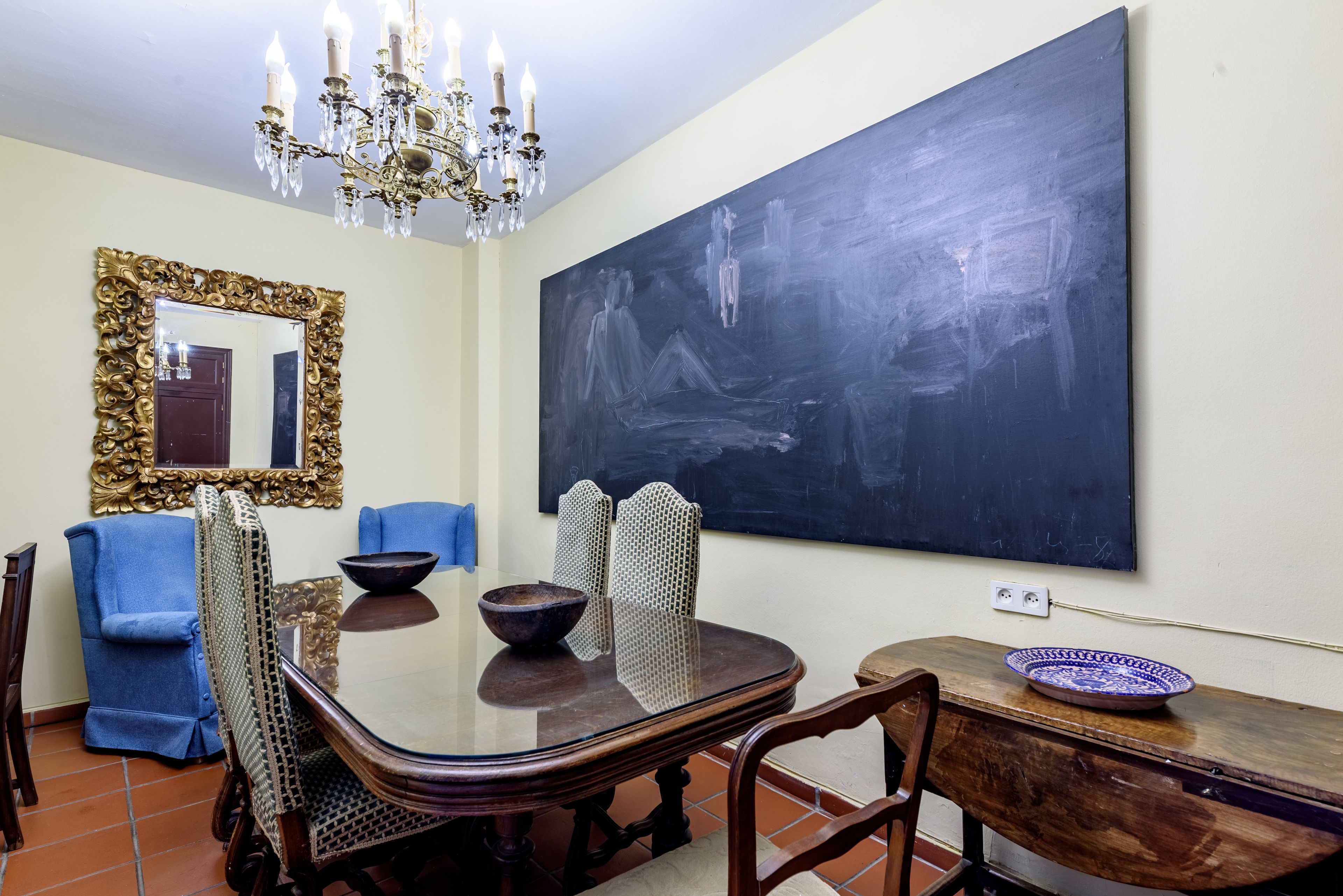 Un comedor con una gran mesa, sillas, una araña, un cuadro abstracto de gran tamaño y un espejo con un marco ornamentado.