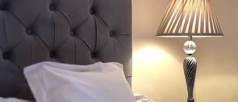 Una cama bien tendida con un almohadón blanco y un cabecero tapizado en gris. Una lámpara de mesa con pantalla texturizada está colocada sobre una mesita de noche junto a la cama.