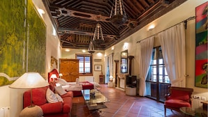 A spacious living room with a red sofa, a glass coffee table, and a fireplace. There is a large window with curtains, a wooden ceiling, and a painting on the wall. - Palacio de Santa Inés hotel (Granada)