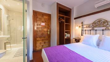 Habitación de hotel con cabina de ducha de vidrio, armario de madera, cama con colcha morada y mesita de noche con lámpara.