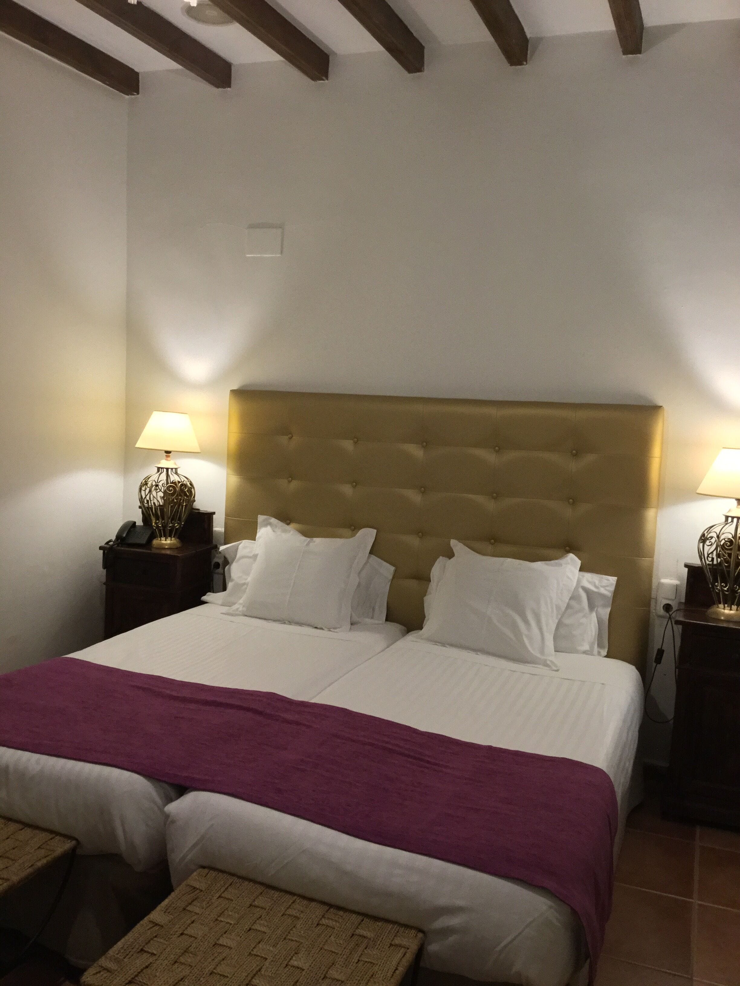 A double bed with a tufted headboard, two bedside tables with lamps, a purple throw blanket, and a woven bench.