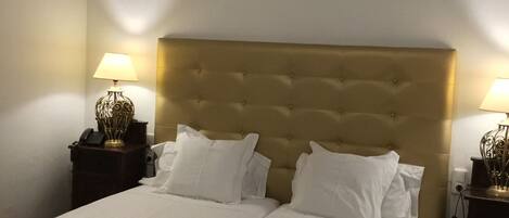 A double bed with a tufted headboard, two bedside tables with lamps, a purple throw blanket, and a woven bench.