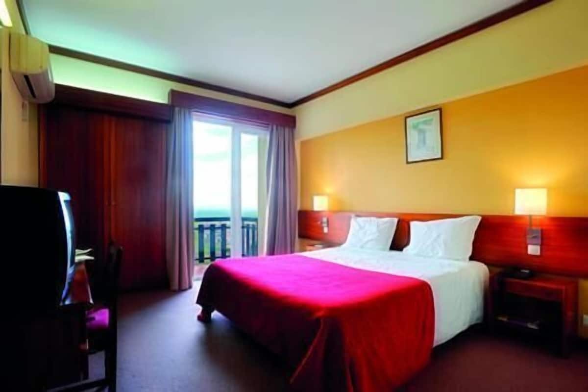 A hotel room with a bed, a desk, a chair, a nightstand, and a window with curtains.
