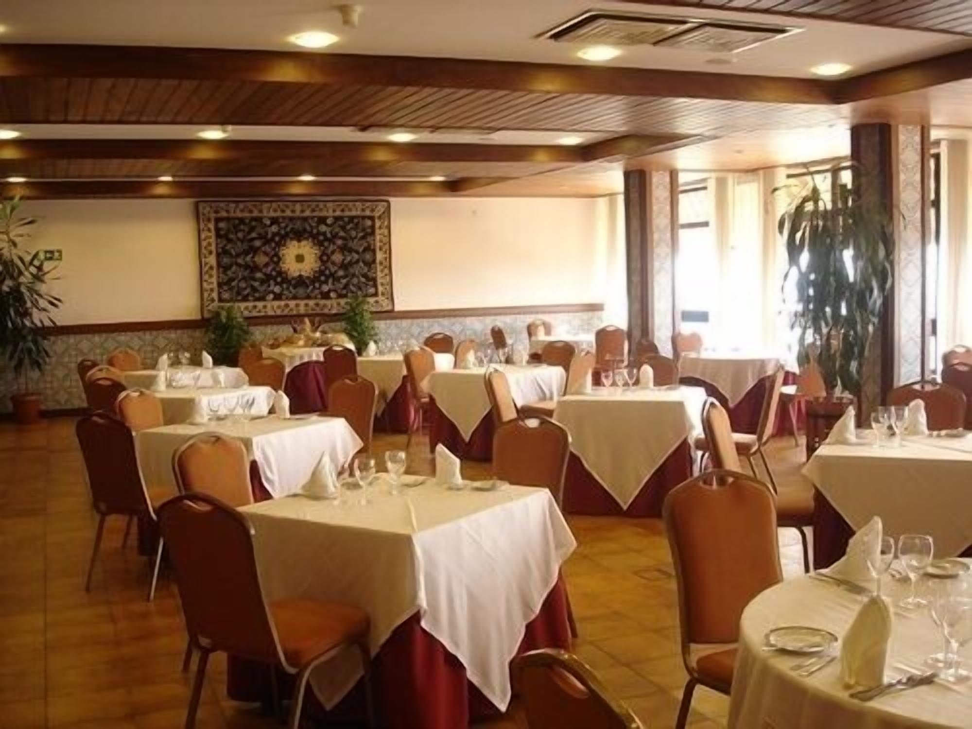 A dining area with multiple round tables covered with white tablecloths, arranged in a spacious room with wooden flooring and a decorative wall hanging.