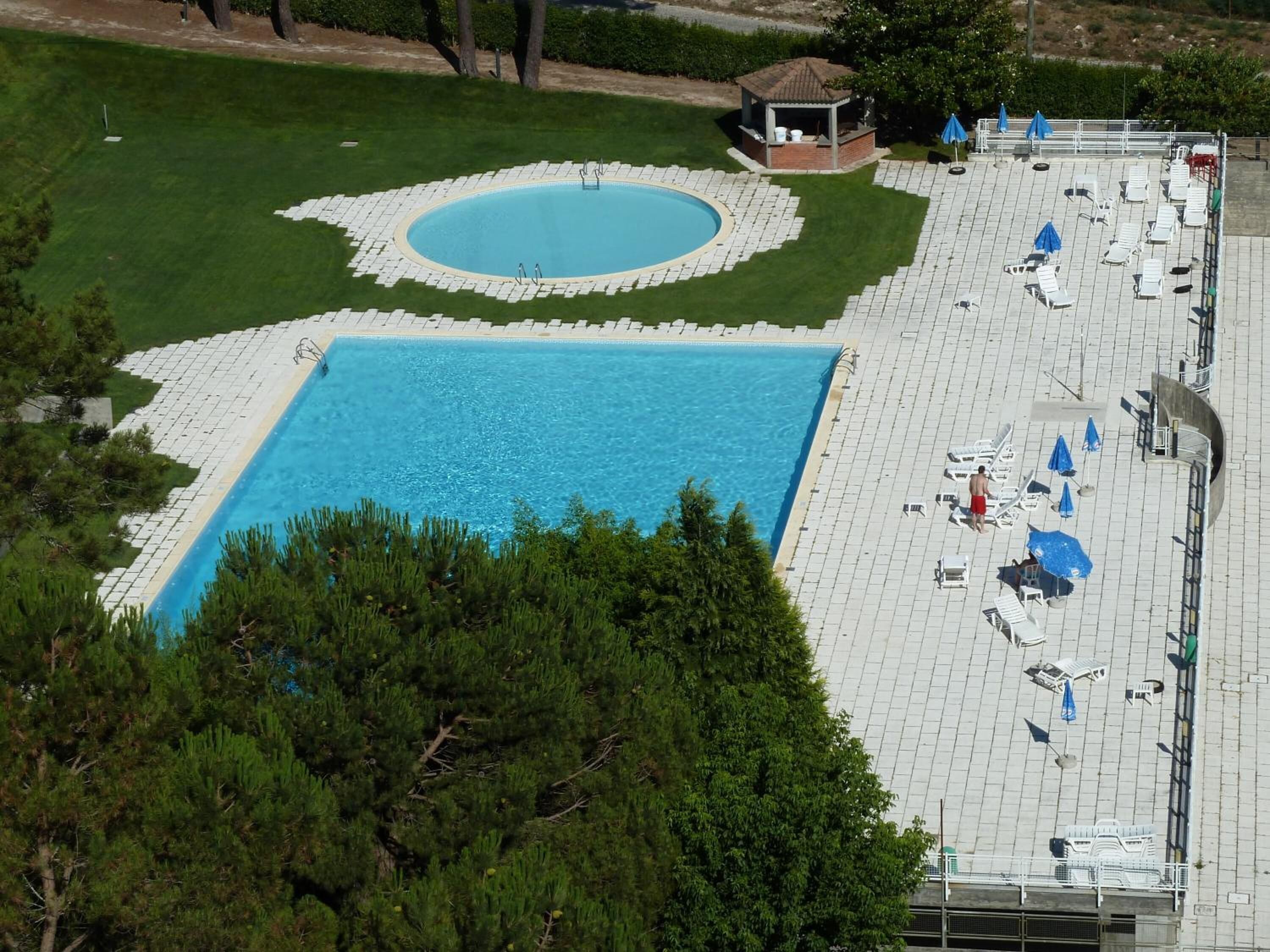 A large rectangular pool with a smaller circular pool, surrounded by a paved deck with lounge chairs and umbrellas.