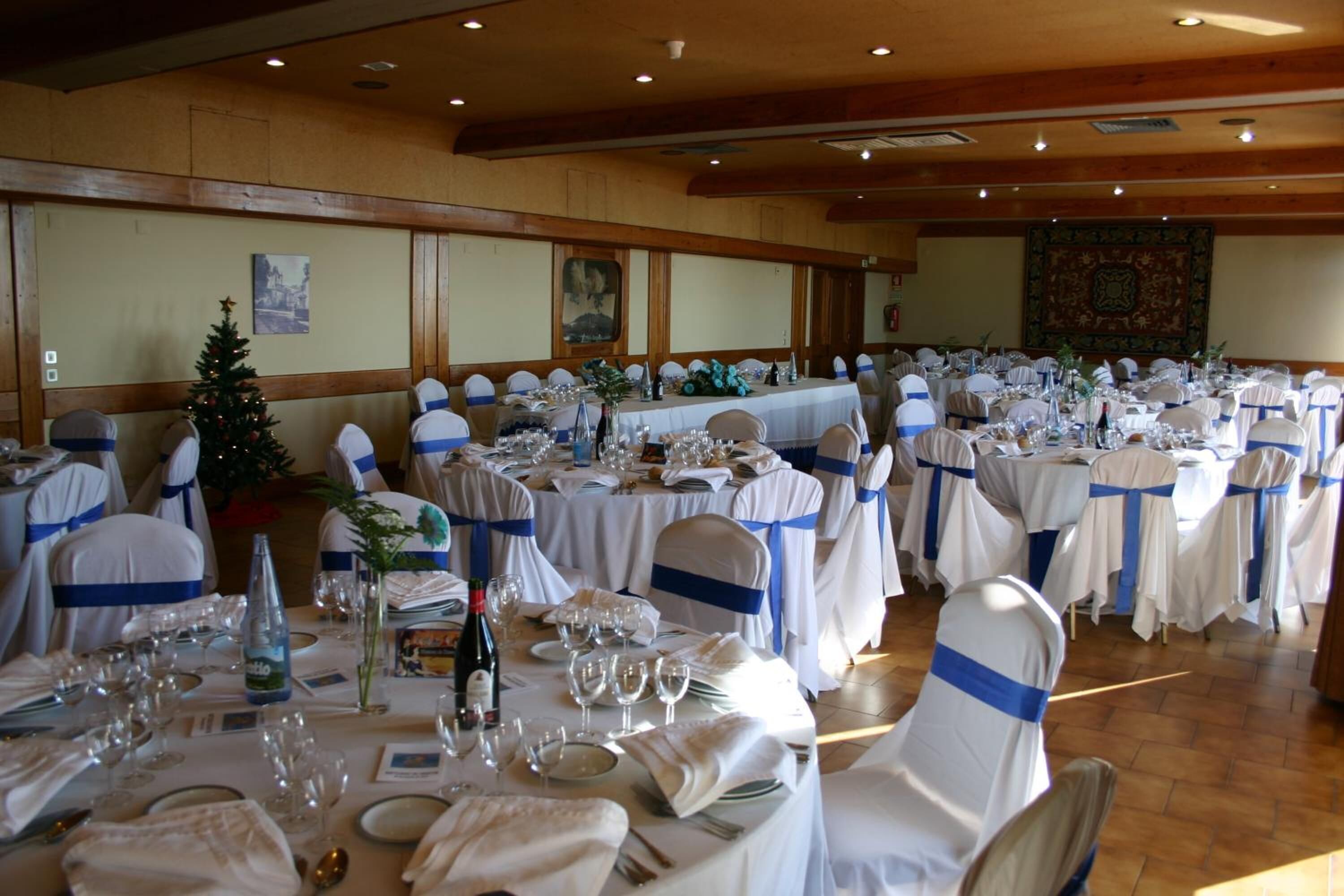 Un salón de banquetes con mesas redondas dispuestas para un evento formal, con manteles blancos y cintas azules en las sillas.