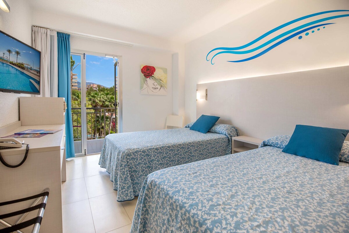 Habitación de hotel con dos camas, un escritorio, una silla, un cuadro y vista a una piscina y palmeras.