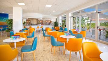A spacious indoor area with round tables and chairs, a bar area in the background, and large windows providing views of the outdoors.