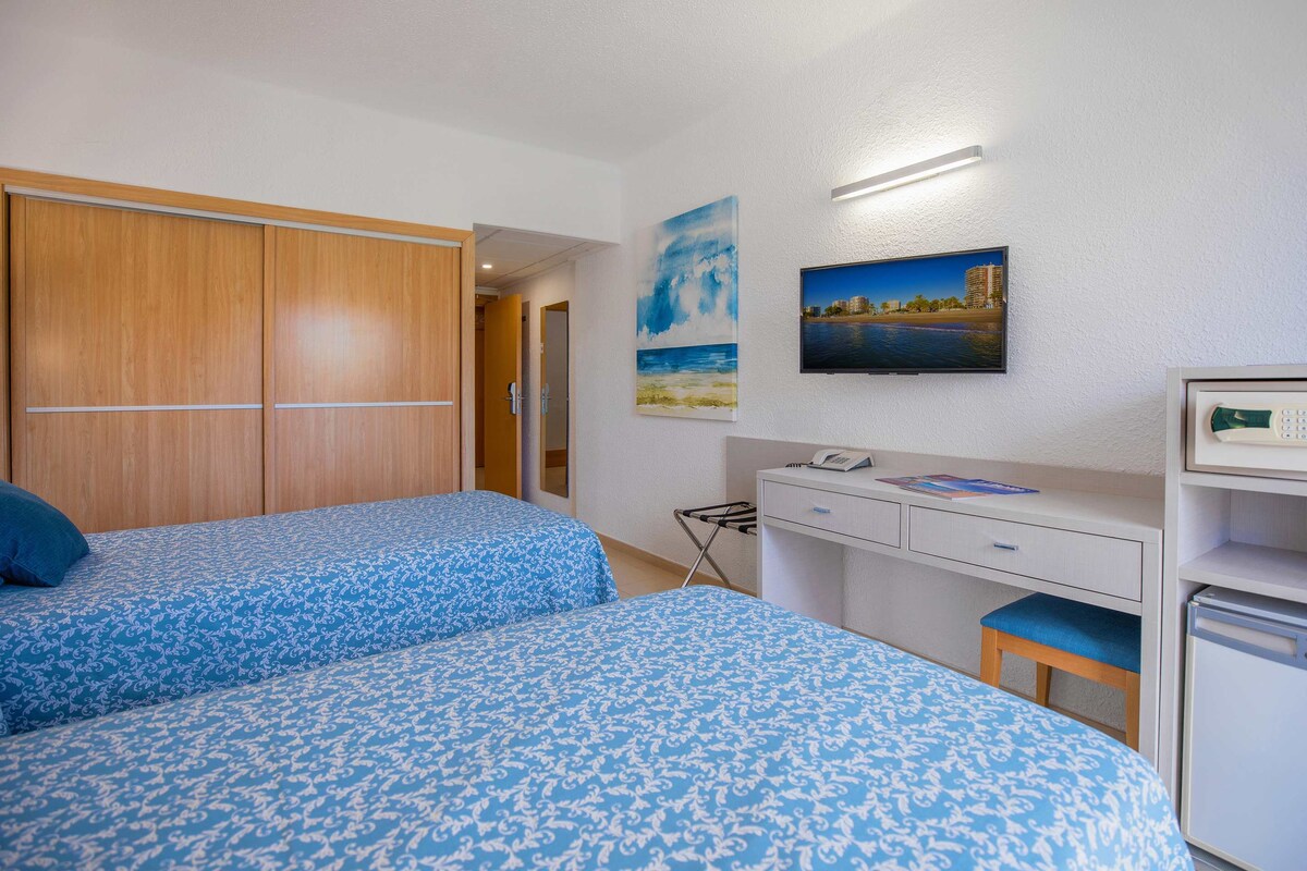A room with two beds, a desk, a chair, a small refrigerator, and a wall-mounted TV.