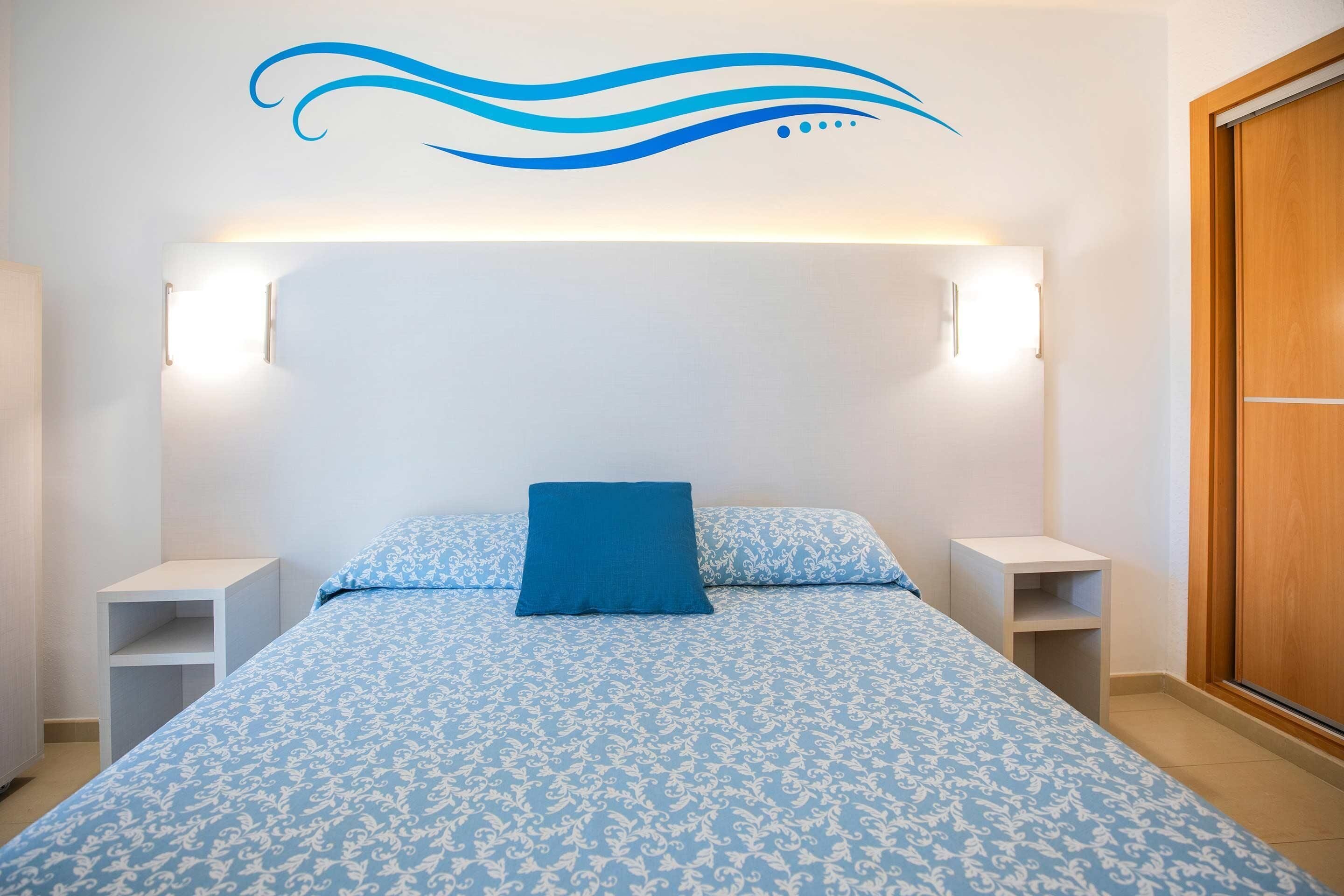 Un dormitorio con cama, una colcha azul estampada, almohada azul, dos mesitas de noche y un adorno en la pared.