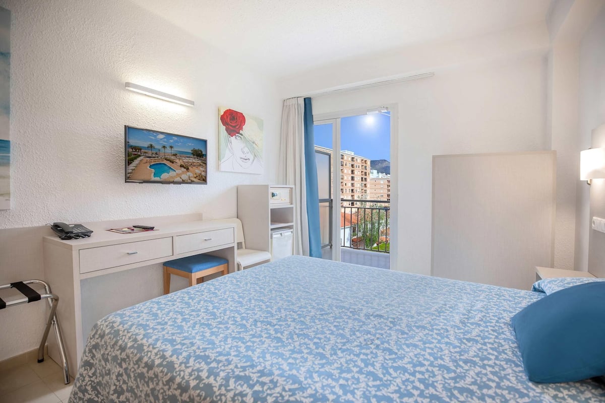 A hotel room with a bed, desk, chair, and a view of the city through a window.