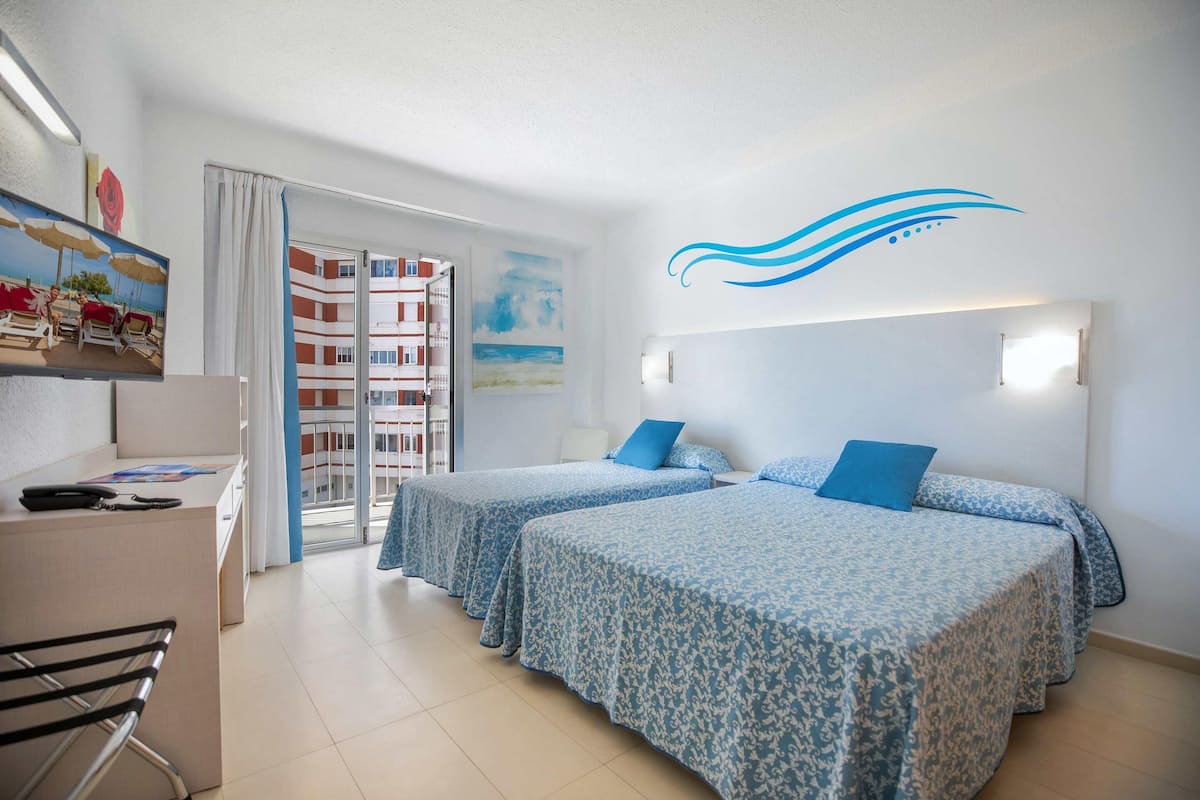 A hotel room with two beds, a desk, a chair, and a view of a building with balconies.