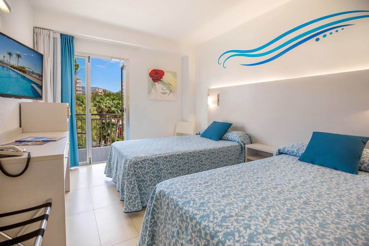 A hotel room with two beds, a desk, a chair, a painting, and a view of a pool and palm trees.