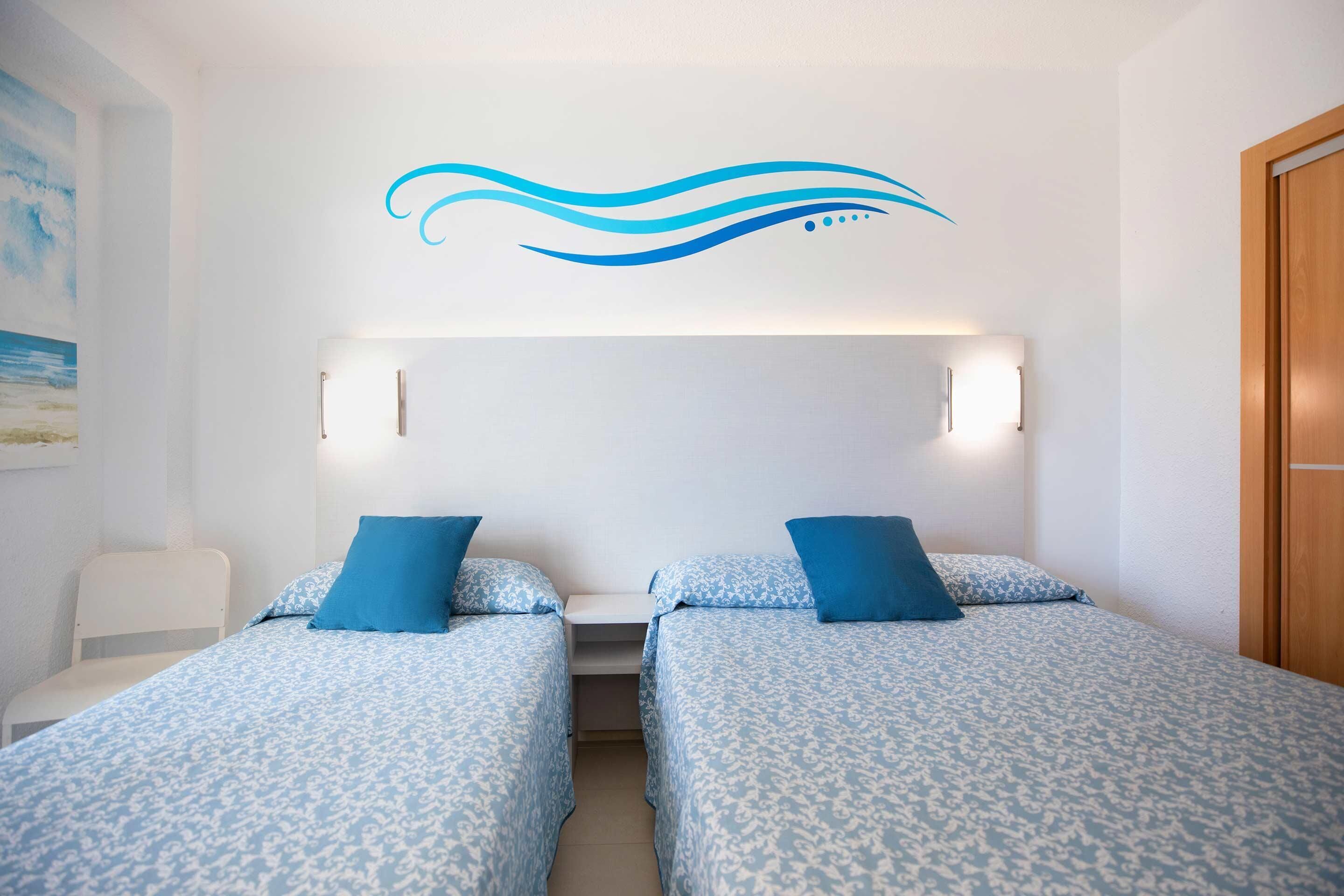 A hotel room with two beds, each with a blue patterned bedspread and matching pillows. There is a white chair, a small table, and a wall-mounted light fixture above the beds.