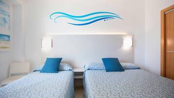 A hotel room with two beds, each with a blue patterned bedspread and matching pillows. There is a white chair, a small table, and a wall-mounted light fixture above the beds.
