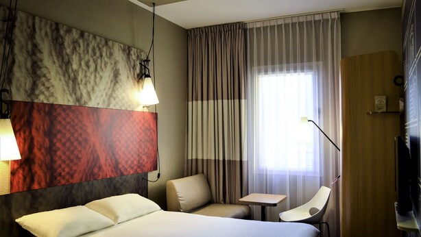 Ein Hotelzimmer mit Bett, Schreibtisch, Stuhl, Lampe und einem großen Fenster mit Vorhängen.