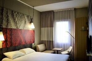 Ein Hotelzimmer mit Bett, Schreibtisch, Stuhl, Lampe und einem großen Fenster mit Vorhängen.