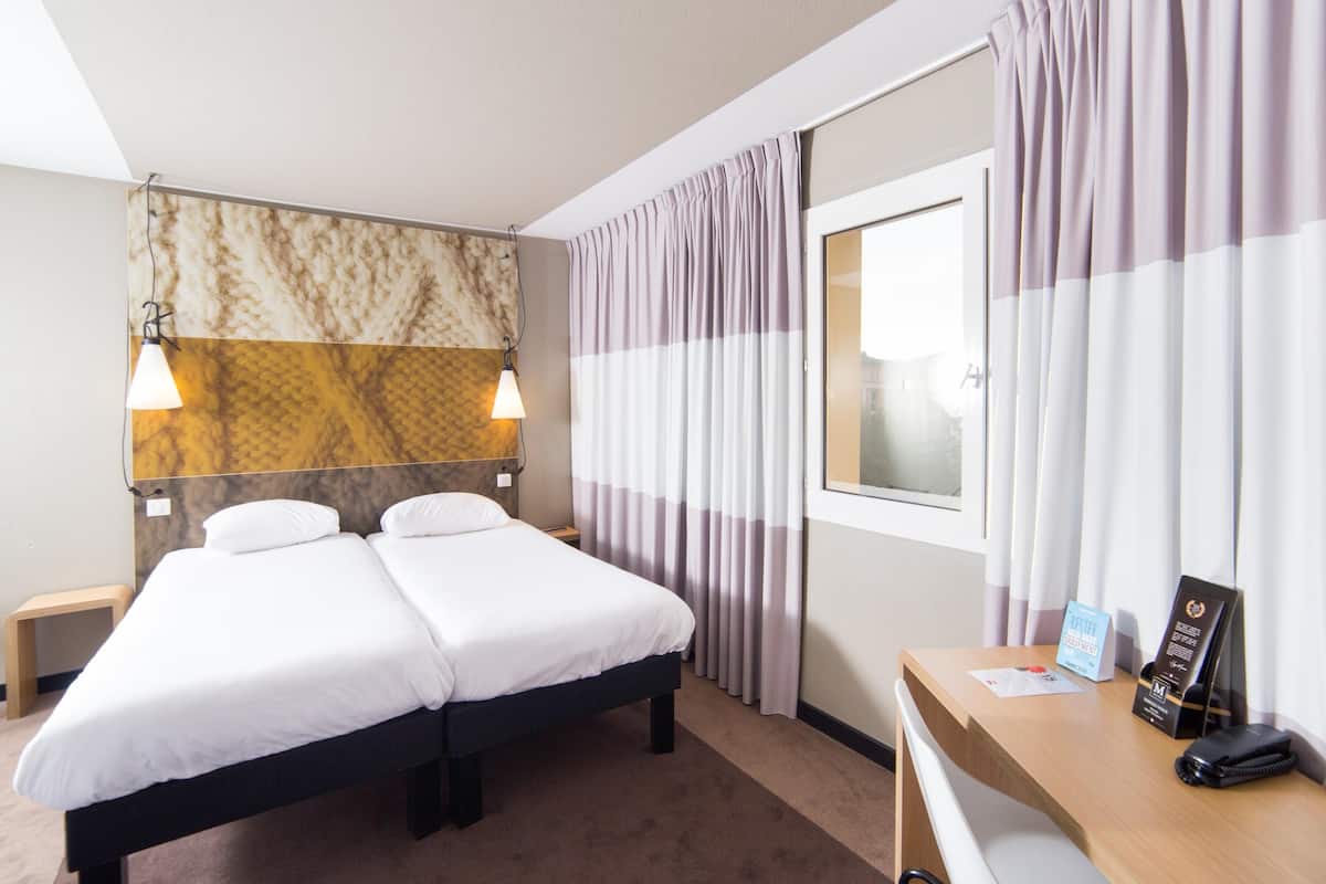 Une chambre d’hôtel avec un lit double, deux tables de chevet, un bureau avec une chaise, une fenêtre avec des rideaux et une tête de lit à motifs de marbre.