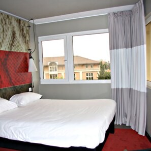 Een hotelkamer met een bed, een raam met gordijnen en uitzicht op gebouwen.